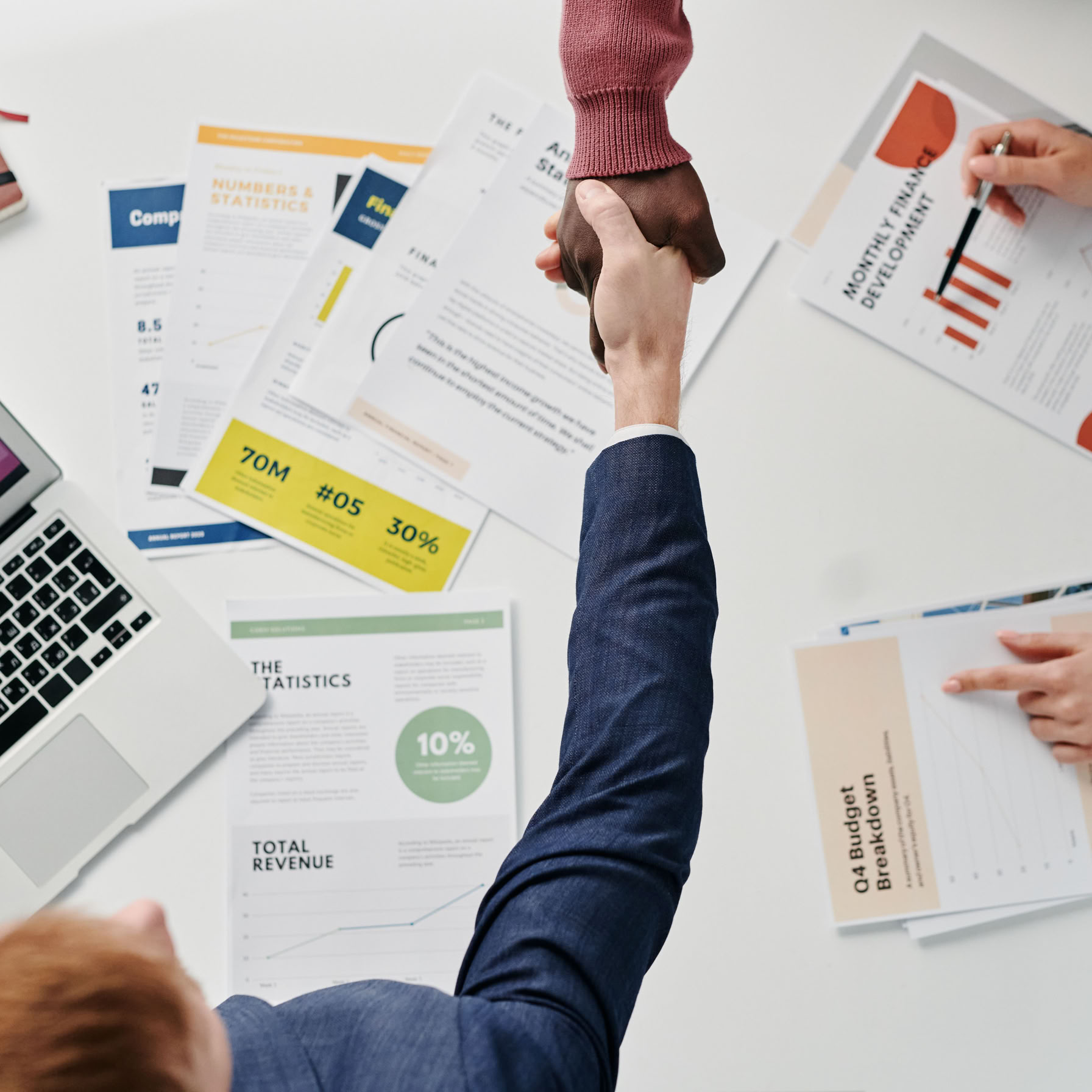 Business professionals engaging in a handshake over a desk filled with financial documents and a laptop.