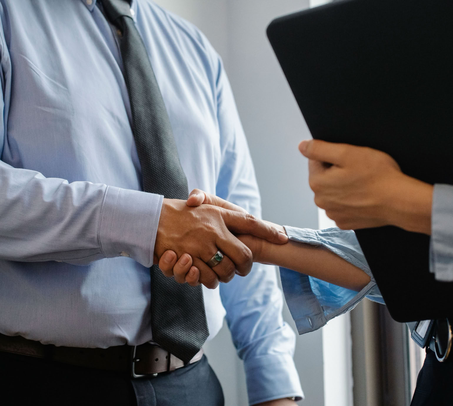 Crop anonymous man in formal shirt and tie shaking hand of woman with black folder while meeting in office