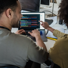 Two programmers working together on a laptop, discussing code in a modern office setting.