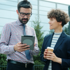 Two professionals discuss business strategies using a tablet outside a modern building.